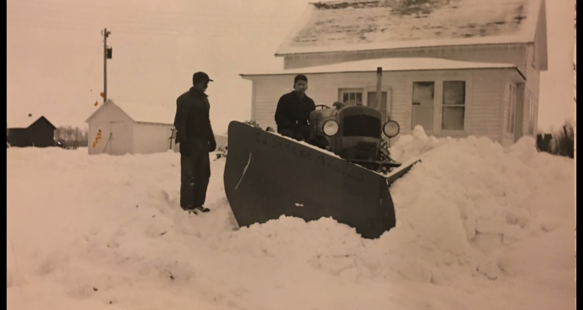 Two men with a homemade V-plow clearing deep snow in front of the farmhouse